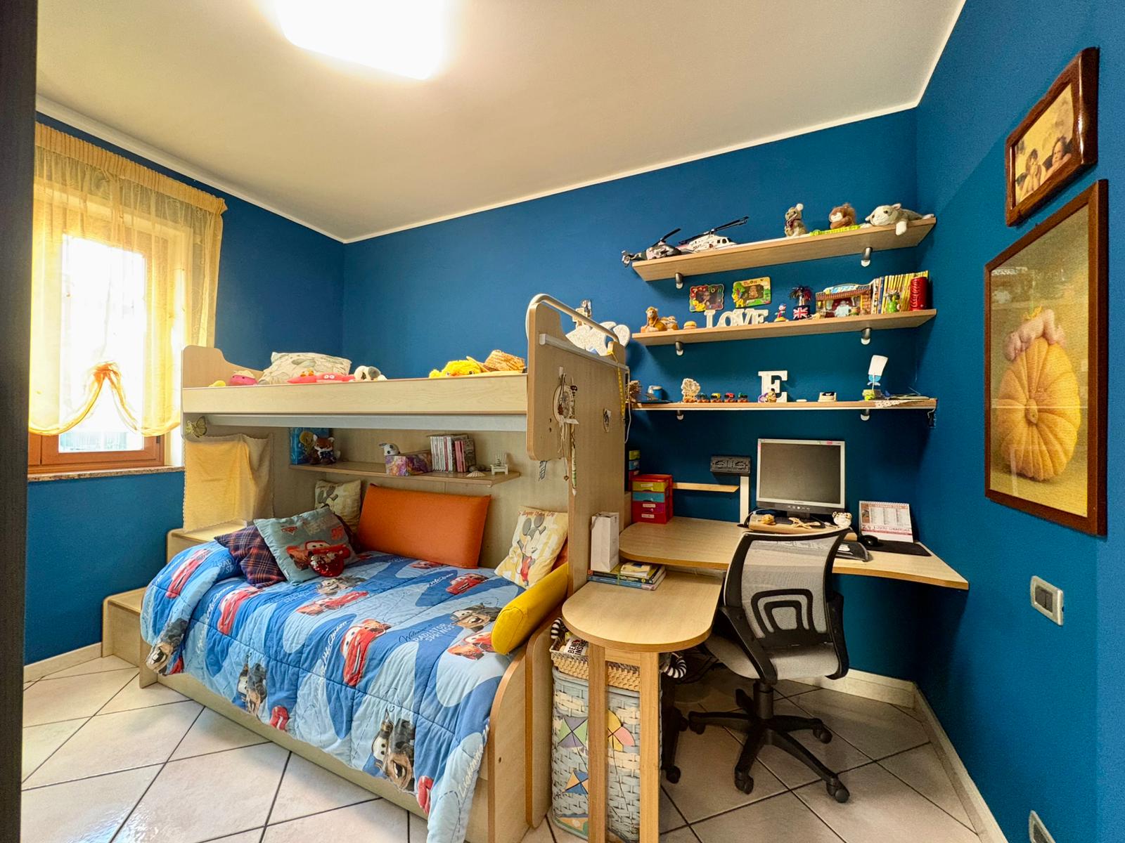 Letto a castello con scrivania e scaffali per camera bambini, pareti blu, arredamento funzionale, decorazioni colorate, ambiente accogliente, ideale per ragazzi, zona studio e riposo, casa completa.