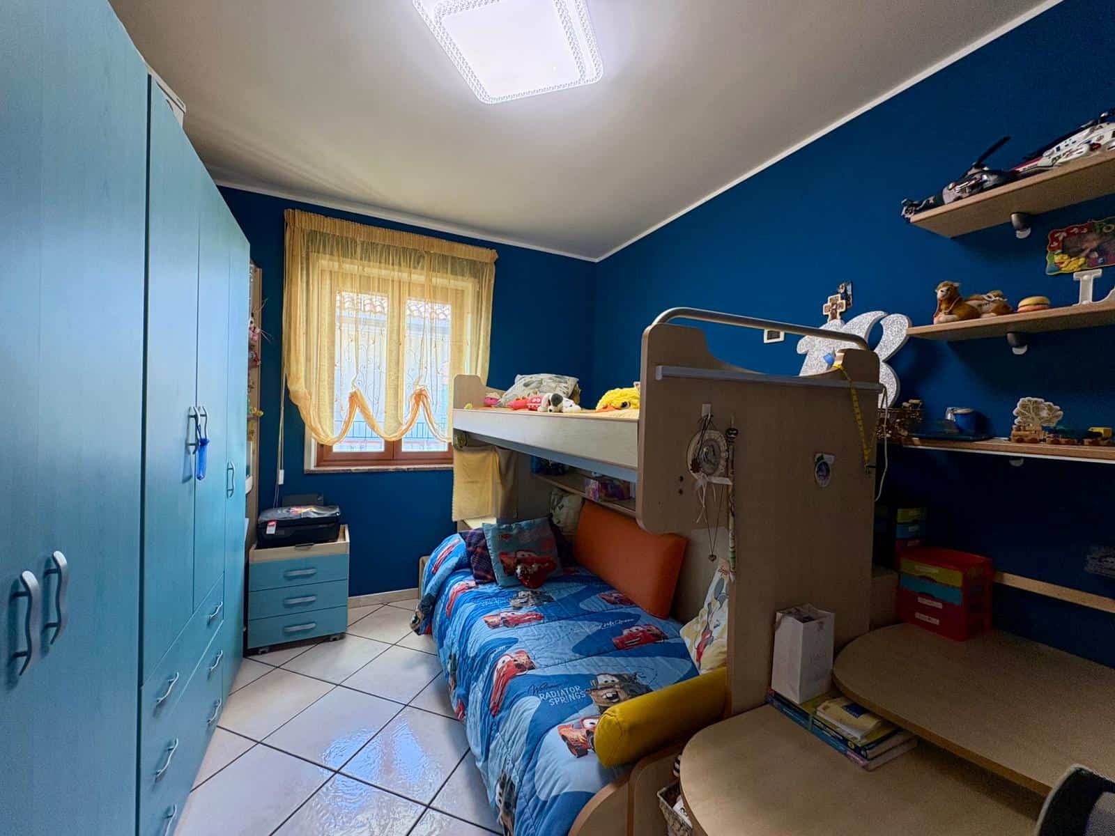 Letto a castello per bambini, stanza con pareti blu, arredi in legno chiaro, armadio, tende gialle, decorazioni per bambini, giocattoli e libri, luce naturale, ambiente accogliente e funzionale per la cameretta.
