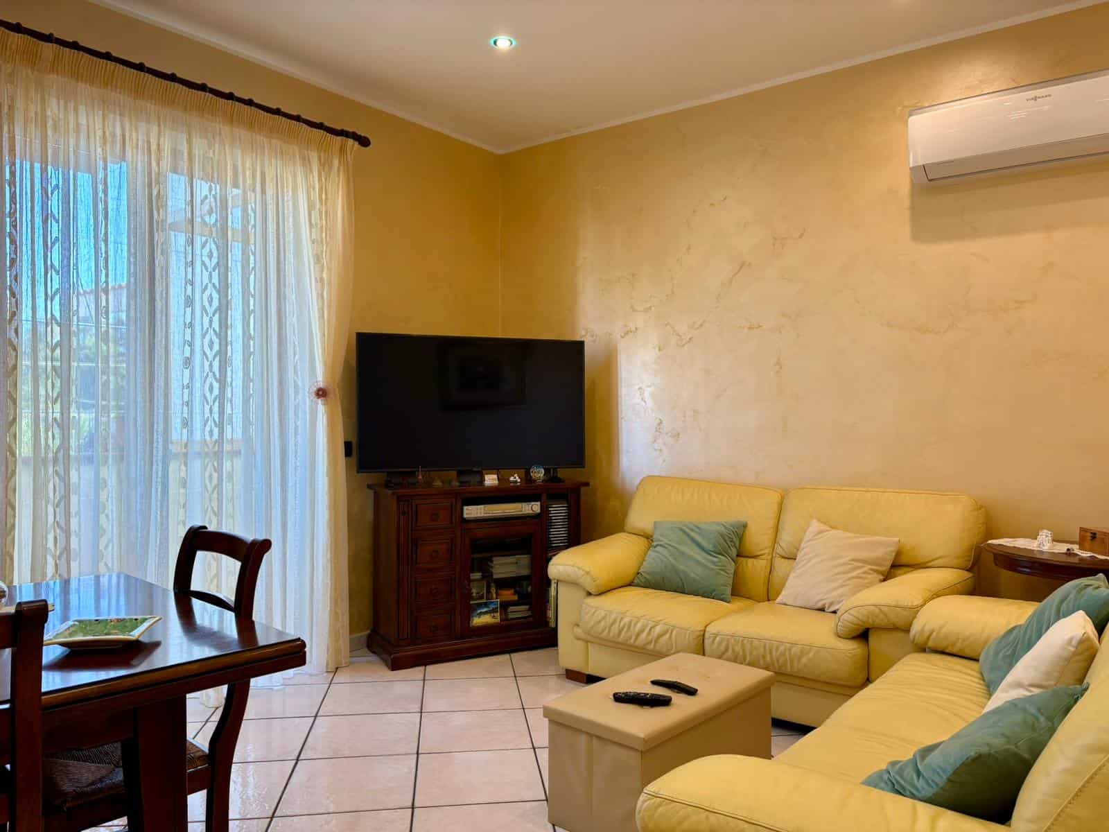Comfortable soggiorno con divani gialli, TV e ottima illuminazione naturale dalle tende sheer, ambiente accogliente con stile classico e ventilato per momenti di relax.