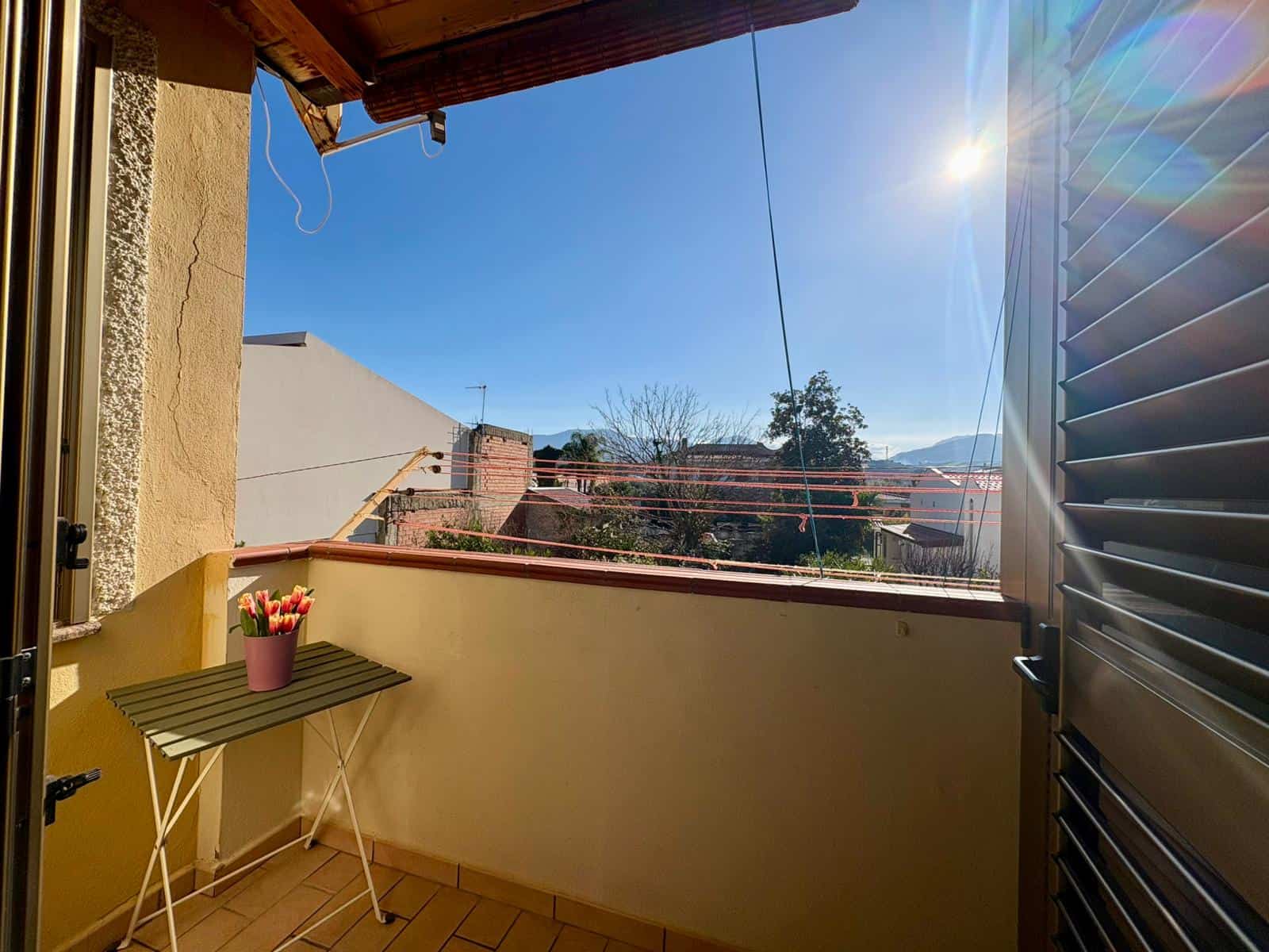 Vista balcone soleggiato con vasi di tulipani, tettoie e linee di lavatrici all'aperto, scenario di case del centro storico, con montagne sullo sfondo, cielo azzurro e sole splendente, ideale soluzione Immobiliare NextCasa.