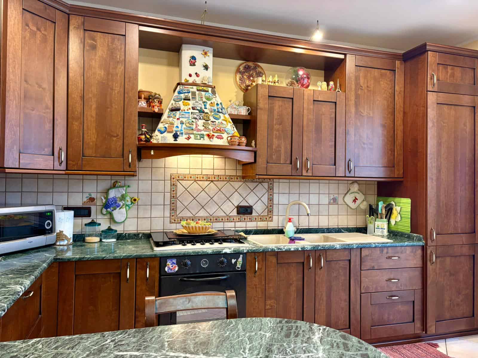 Cucina in stile classico con ante in legno scuro, piano in marmo verde, e decorazioni colorate, ideale per vivere momenti conviviali e americani, con elettrodomestici e dettagli accoglienti.