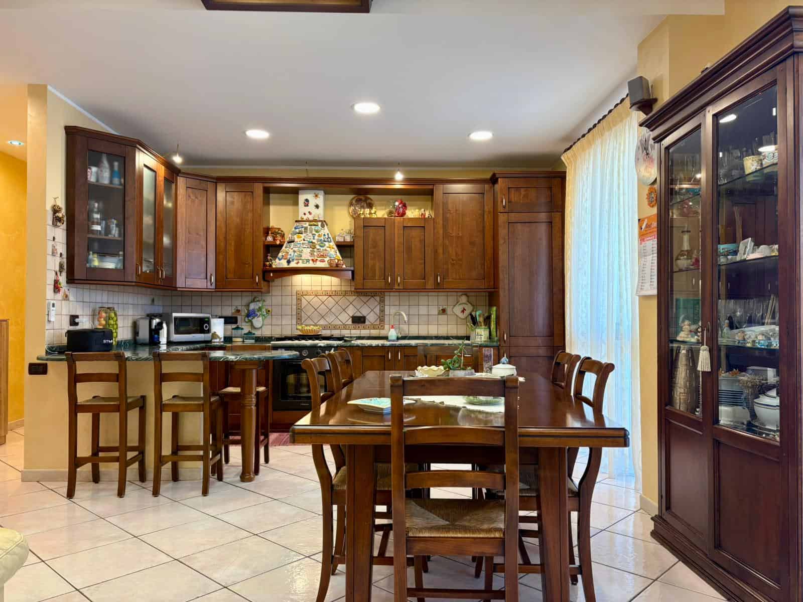 1. Cucina spaziosa in stile classico con mobili in legno scuro, piano cottura a gas e zona pranzo con tavolo e sedie in legno, illuminata da luce naturale e luci a soffitto.