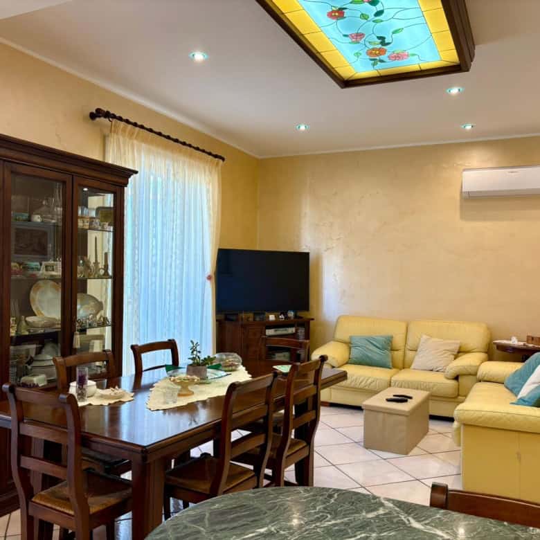 Salotto con divani gialli, tavolo da pranzo in legno, credenza in legno scuro, TV LCD, lampadario in vetro colorato, pareti beige, tende leggere, decorazioni eleganti, ambiente accogliente e ben arredato.