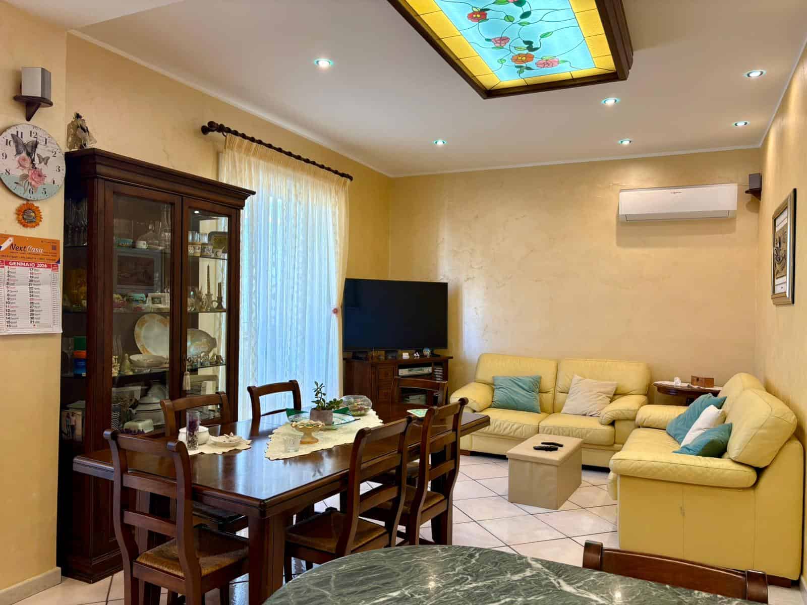 Salotto con divani gialli, tavolo da pranzo in legno, credenza in legno scuro, TV LCD, lampadario in vetro colorato, pareti beige, tende leggere, decorazioni eleganti, ambiente accogliente e ben arredato.