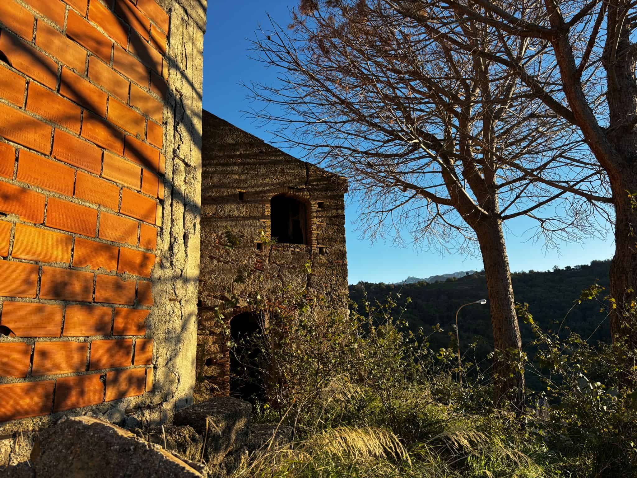 Vecchio edificio in pietra e mattoni in campagna con albero spoglio e vista sulle montagne, ambiente rustico e suggestivo per case di charme e immobili di pregio.