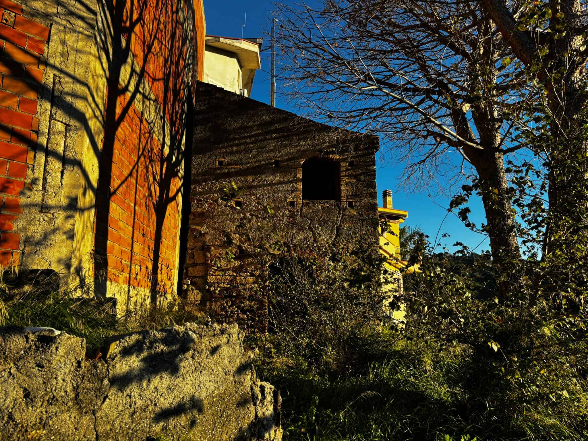Vecchio edificio in pietra con giochi di ombre di alberi, contornato da vegetazione, sotto un cielo blu, atmosfera di charme e rusticità.