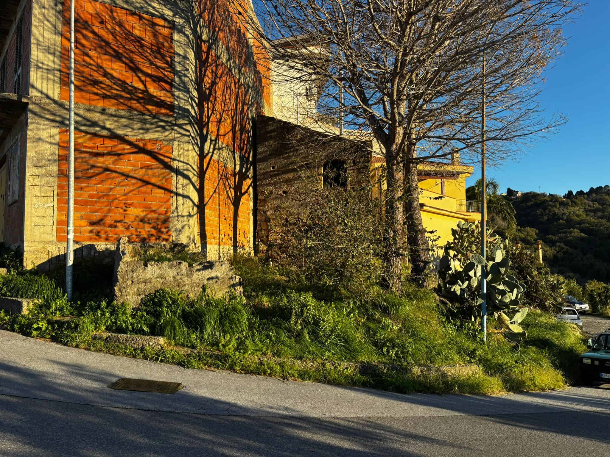 1. Inserto di un edificio in costruzione con pareti di mattoni arancioni e casa gialla sullo sfondo, con alberi e piante verdi lungo la strada, esempio di immobili in zona residenziale di NextCasa.