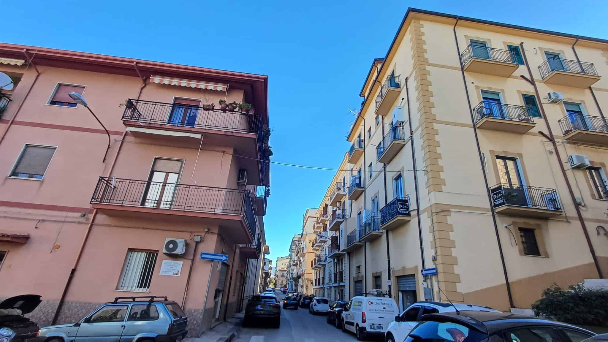 Balcone e facciate di edifici residenziali in un quartiere urbano, con auto parcheggiate lungo la strada e un cielo azzurro, rappresentando l'architettura e la vita quotidiana in città italiane.