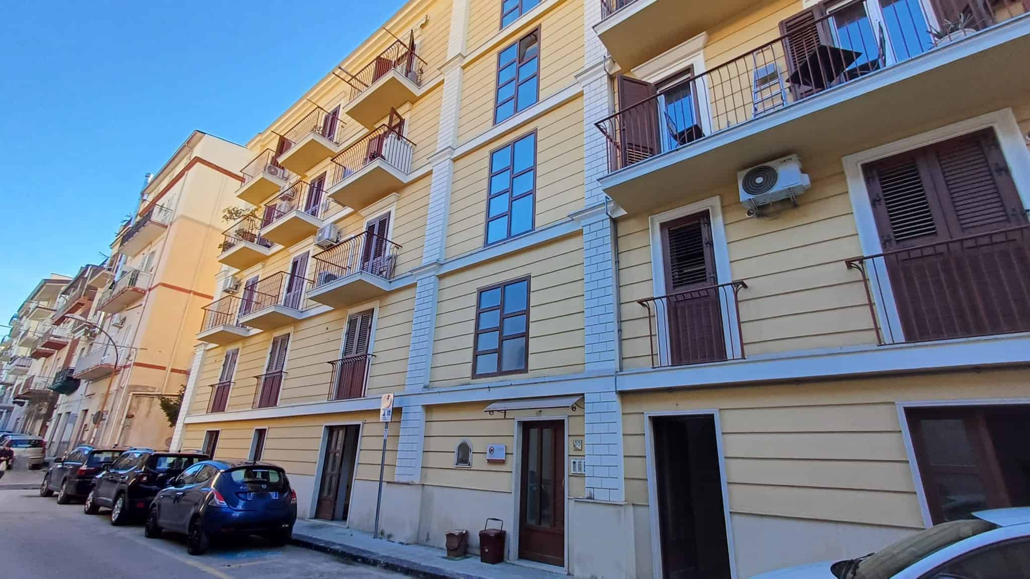 Ampio edificio residenziale con facciata gialla e balconi in ferro battuto, situato in una zona urbana con parcheggi lungo la strada.