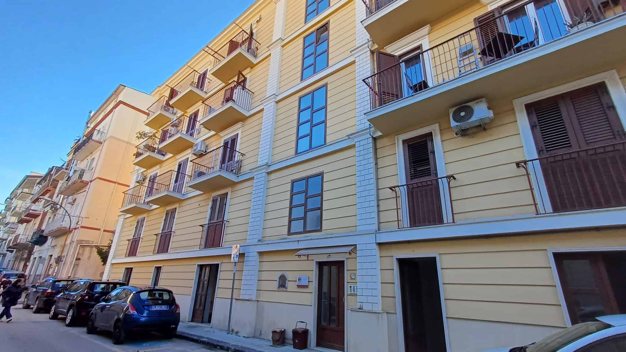 L'immagine mostra un edificio residenziale moderno con balconi e finestre, situato in un'area urbana soleggiata con auto parcheggiate lungo la strada.