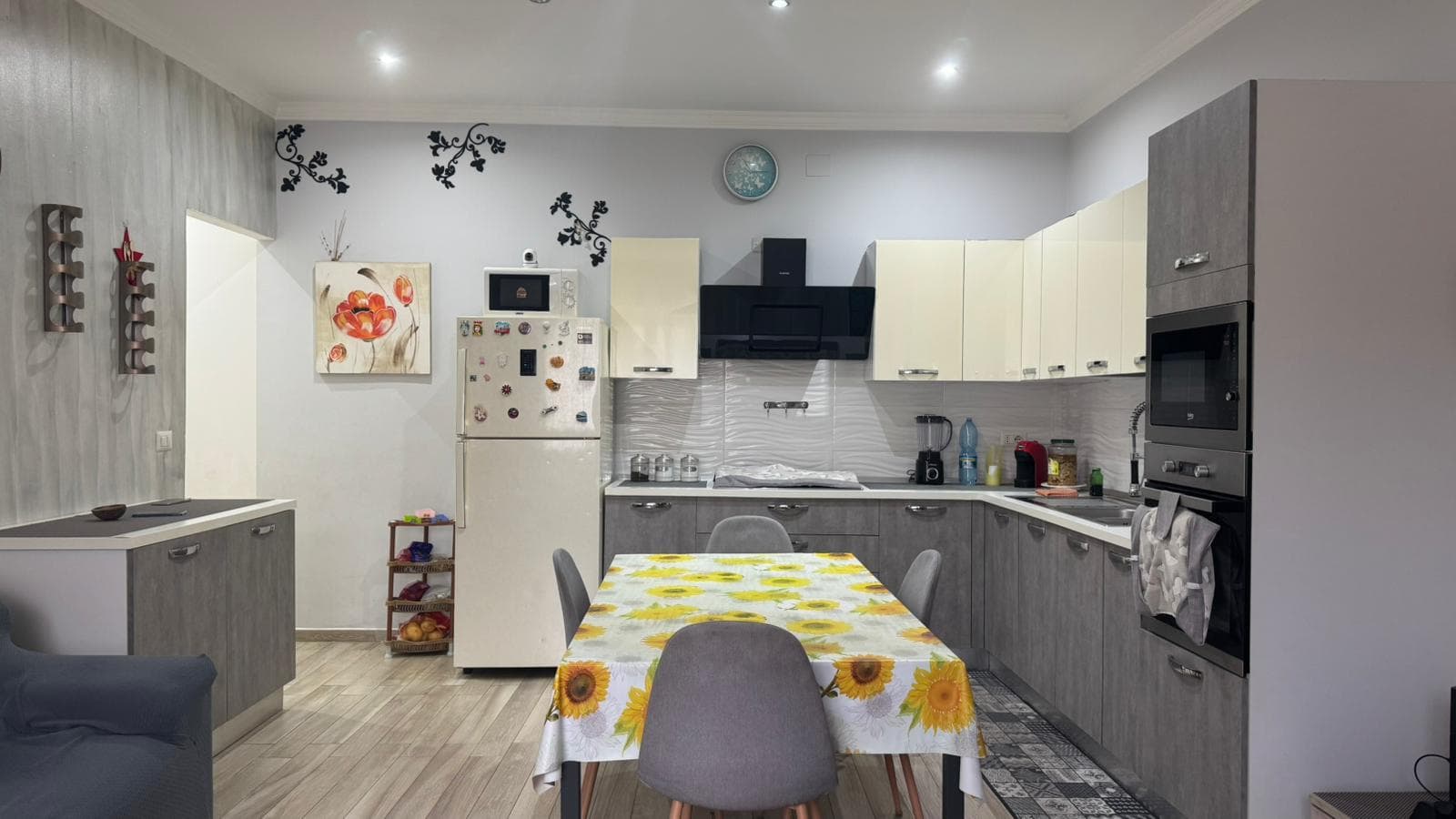 1. Cucina moderna con mobili in legno grigio e bianco, elettrodomestici integrati, tavolo da pranzo con tovaglia a girasoli e decorazioni eleganti, ambiente accogliente e funzionale.