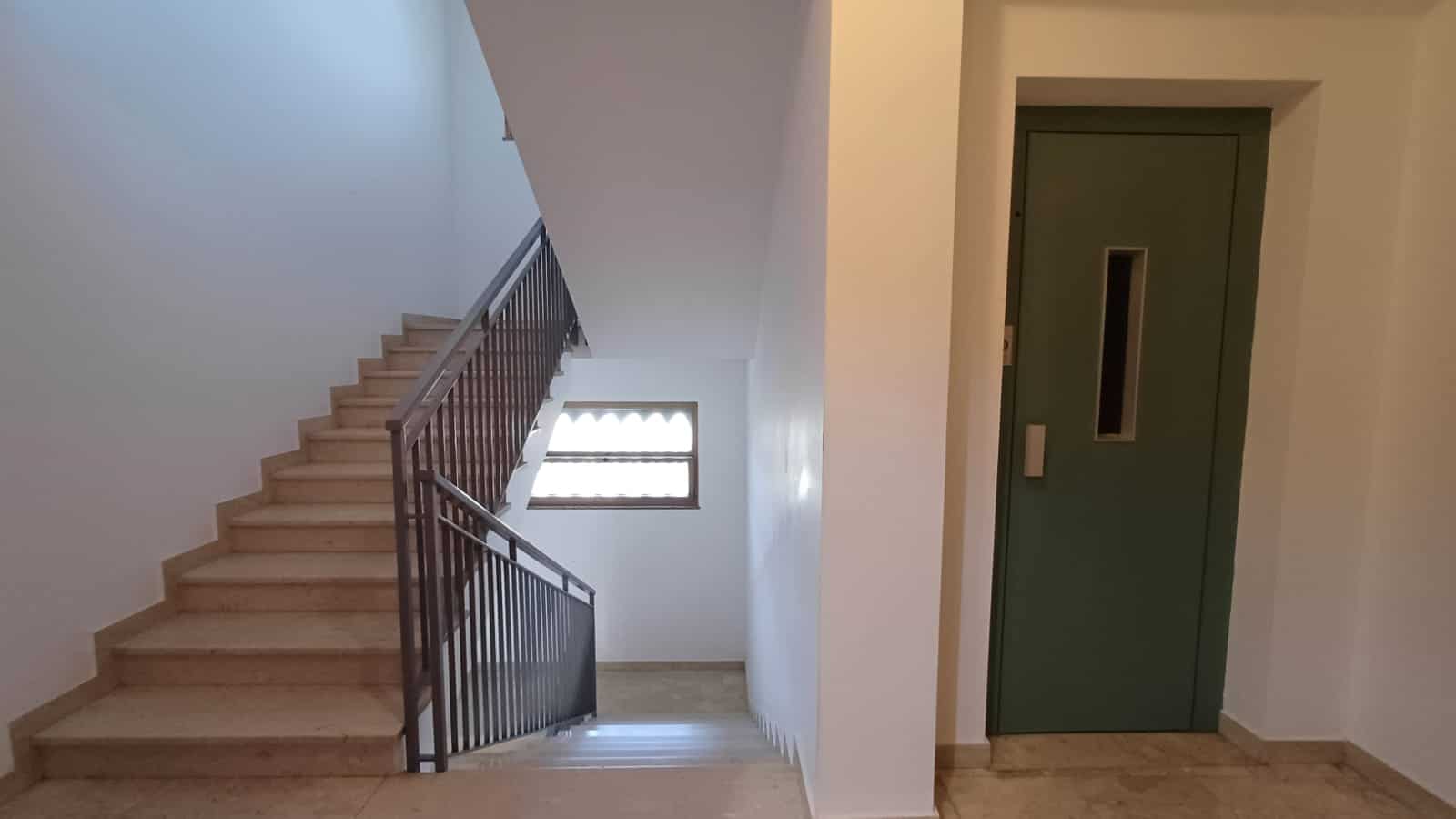 Scendere scale all’interno di un edificio con ascensore e rampa in metallo, pareti bianche e finestra luminosa, ambiente moderno e funzionale per immobili residenziali NextCasa.