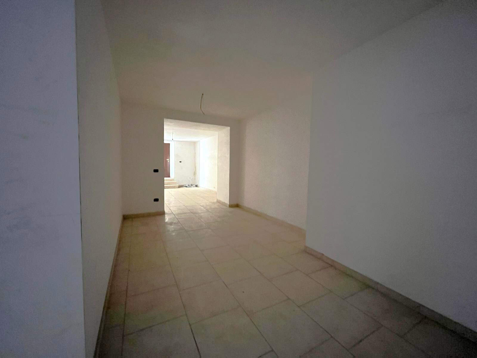 Ampia stanza vuota in fase di ristrutturazione, con pareti bianche e pavimento in piastrelle beige, pronta per essere personalizzata e arredato. Ideale per creare un soggiorno o uno spazio multifunzio.