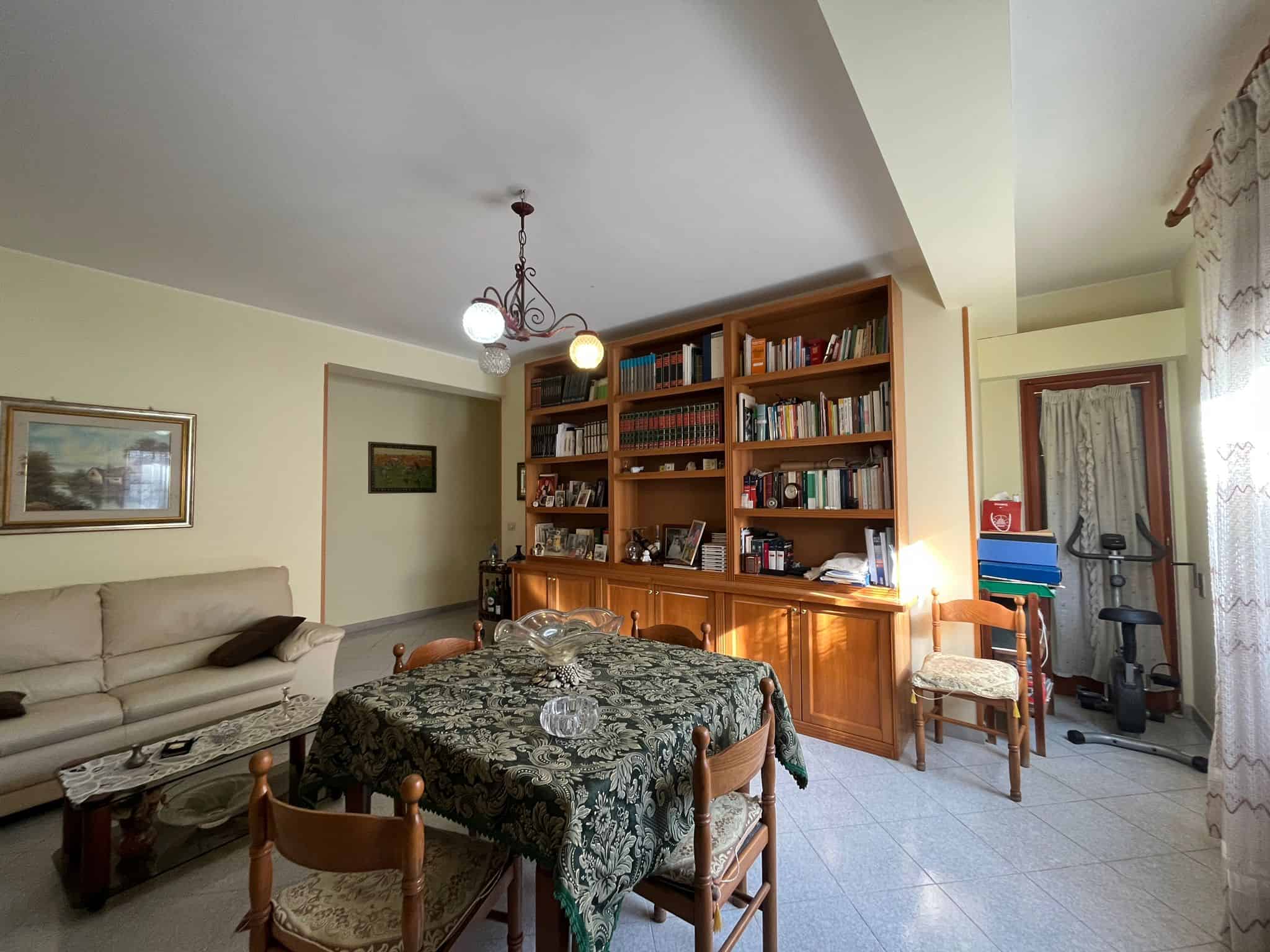 Soggiorno con libreria in legno, divano beige, tavolo con tovaglia decorata, illuminazione a soffitto e zona studio con cyclette, ambiente accogliente e funzionale.