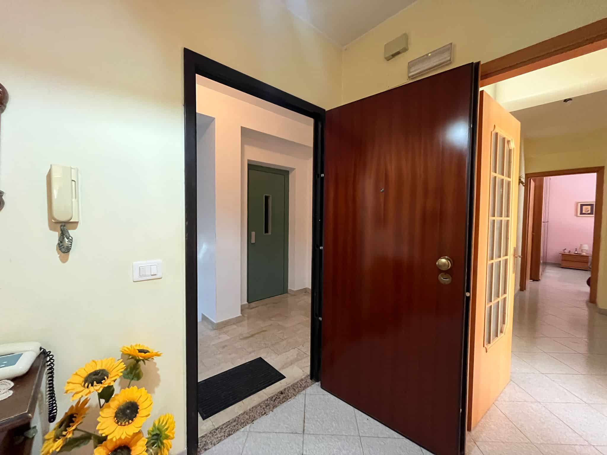 Moderno ingresso condominiale con porta d'ingresso in legno, interfono e pavimento in piastrelle chiara, per un'accoglienza elegante e sicura in appartamenti NextCasa.