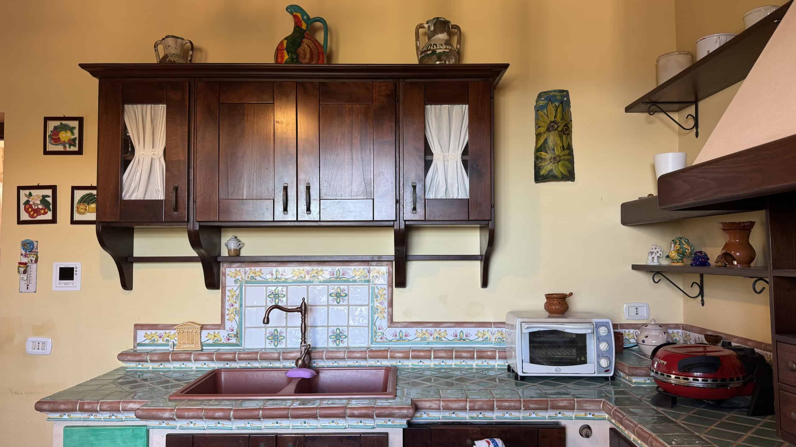 Fornetto di cucina rustico con piani in piastrelle di ceramica, armadietti in legno scuro, decorazioni tradizionali e accessori da cucina, stile campagna italiana, ambientazione accogliente e classica.