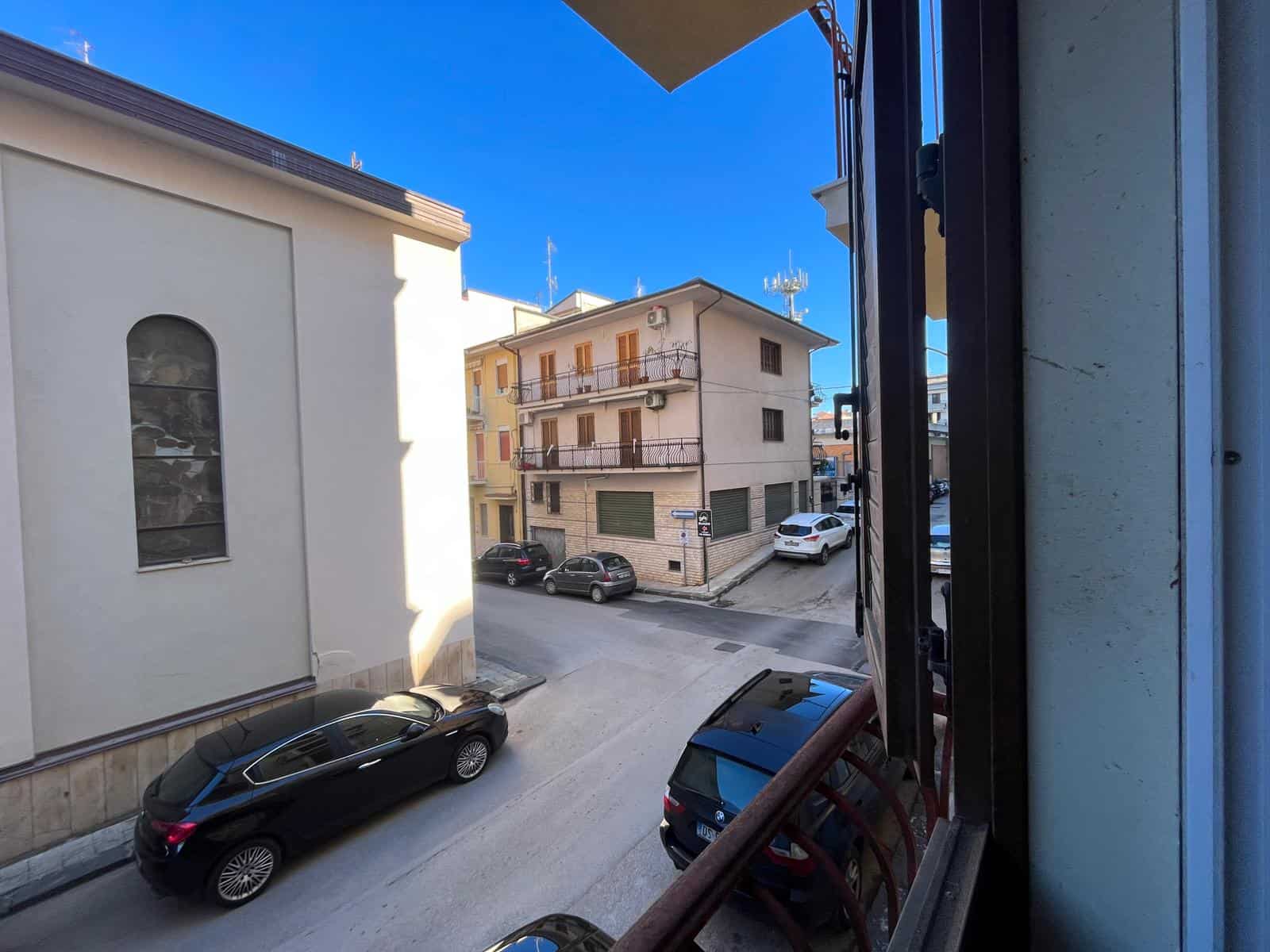 Balcone con vista su strada urbana, edifici residenziali e veicoli parcheggiati, in una giornata soleggiata, rappresentando un tipico scenario di quartiere in Italia.