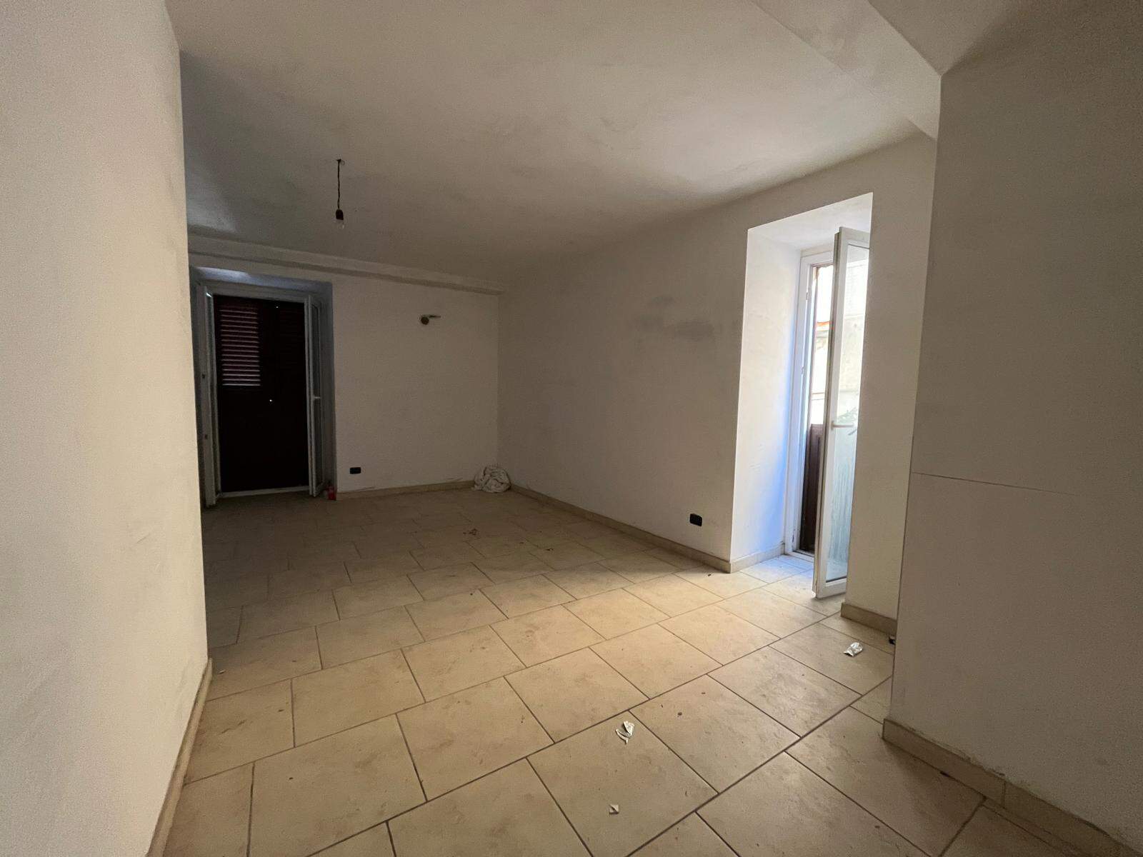 Interno di un appartamento in fase di ristrutturazione con pareti bianche e pavimento in piastrelle beige, luce naturale da una finestra e porta balcone.