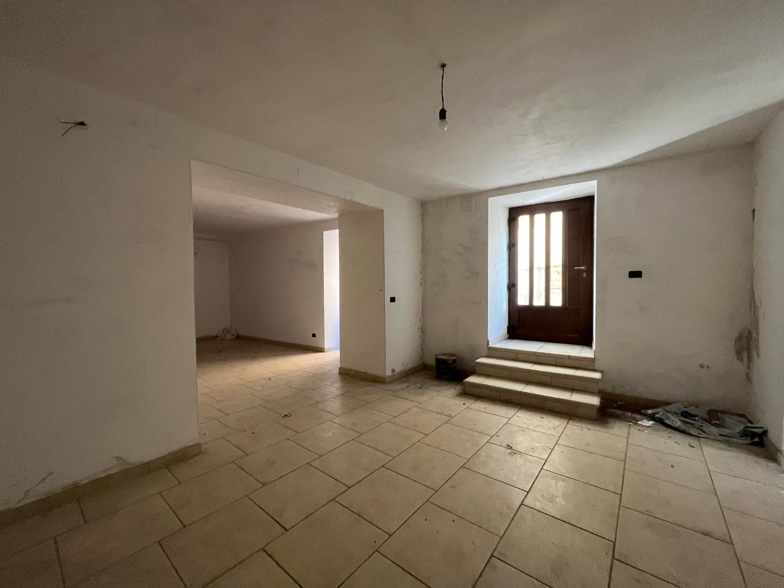 Ampia stanza in fase di ristrutturazione con pareti bianche e pavimento in piastrelle beige, ingresso con porta in legno e scalini, ideale per progetti di ristrutturazione e interior design.