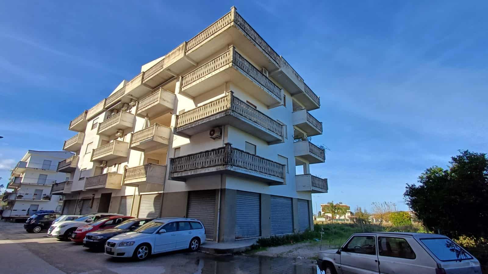 Ampio edificio residenziale con balconi, parcheggio esterno e negozi al piano terra, situato in una zona tranquilla e ben collegata, ideale per investimenti immobiliari e living confortevole in Italia.
