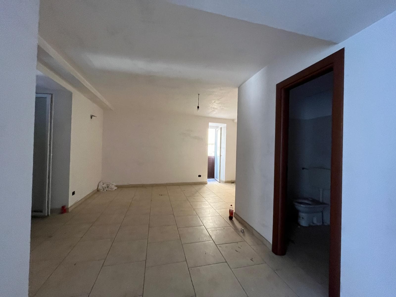 Ampia zona soggiorno open space con pareti bianche e pavimento in piastrelle beige, luminosa grazie alla porta finestra che conduce a un balcone, ideale per ristrutturazione e arredo moderno.