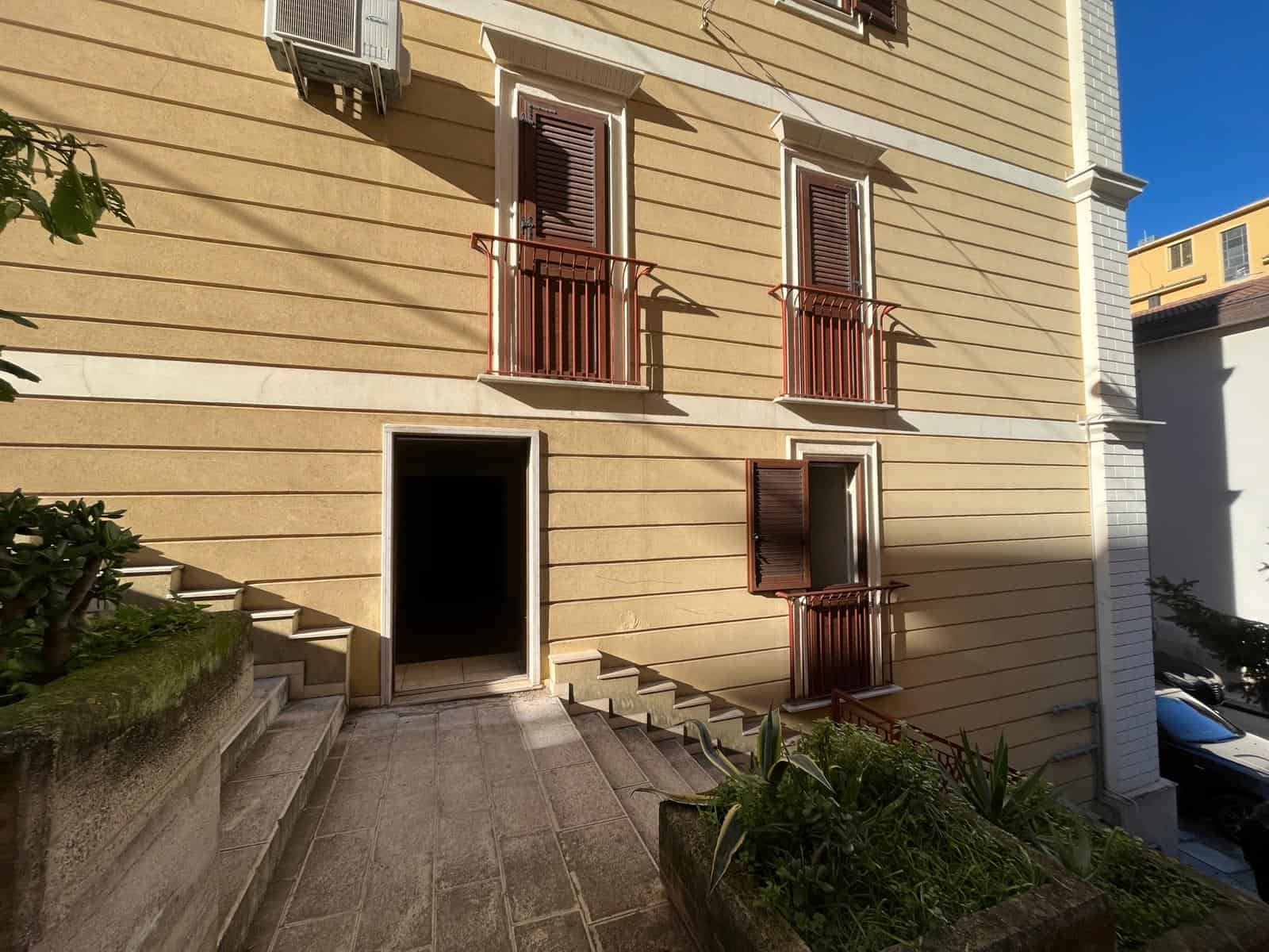 Porte e finestre in legno con balconi in ferro battuto su facciata di edificio residenziale, ingresso principale con scale in pietra, ambiente esterno ben curato e luminoso.
