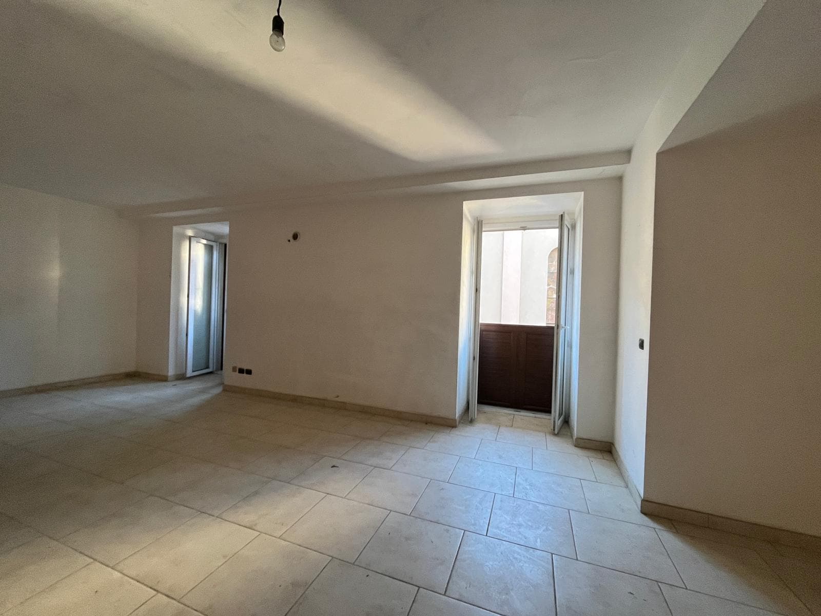 Interno di appartamento vuoto con grandi finestre e porta finestra che dà su un balcone, pavimento in piastrelle chiare, pareti bianche, pronto per arredare.