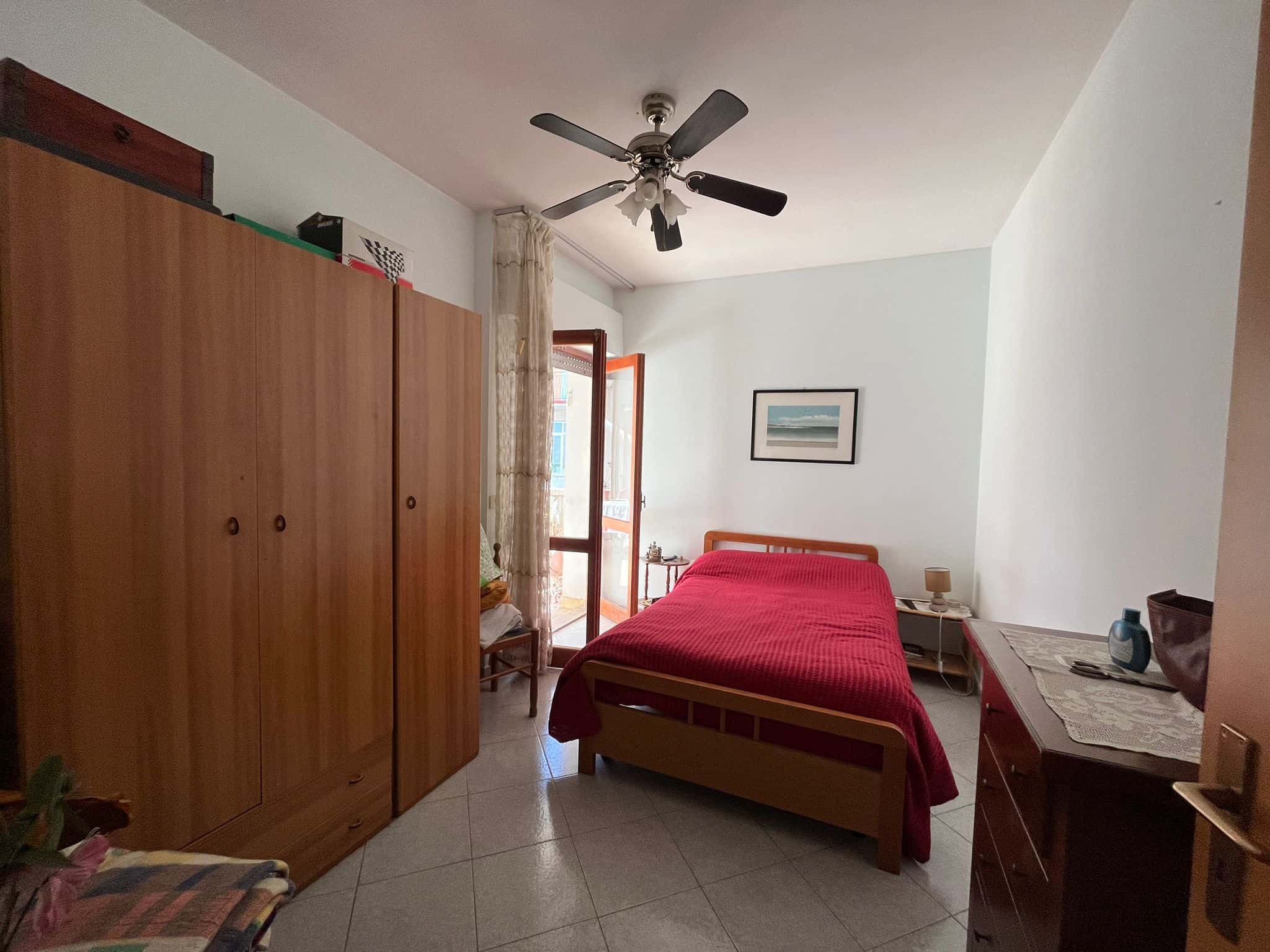 Armadio in camera da letto con balcone, arredamento classico, letto matrimoniale con copriletto rosso, parete con quadro decorativo, oggetti di biancheria e accessori, luce naturale e ventilatore a soffitto.