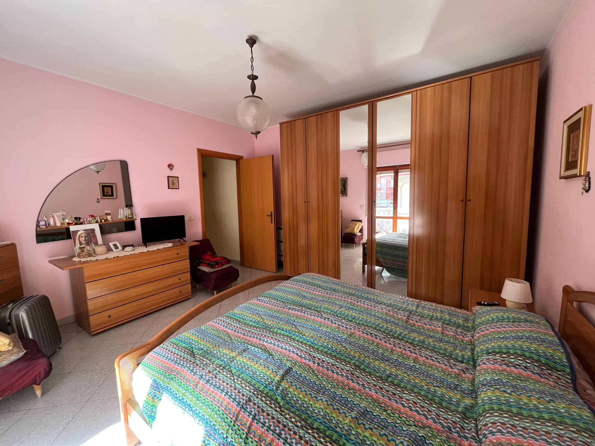 Ampio e luminoso camera matrimoniale con pareti rosa, armadio a parete con specchi, letto con copriletto multicolore, comodino con lampada e specchiera, arredamenti in stile classico.