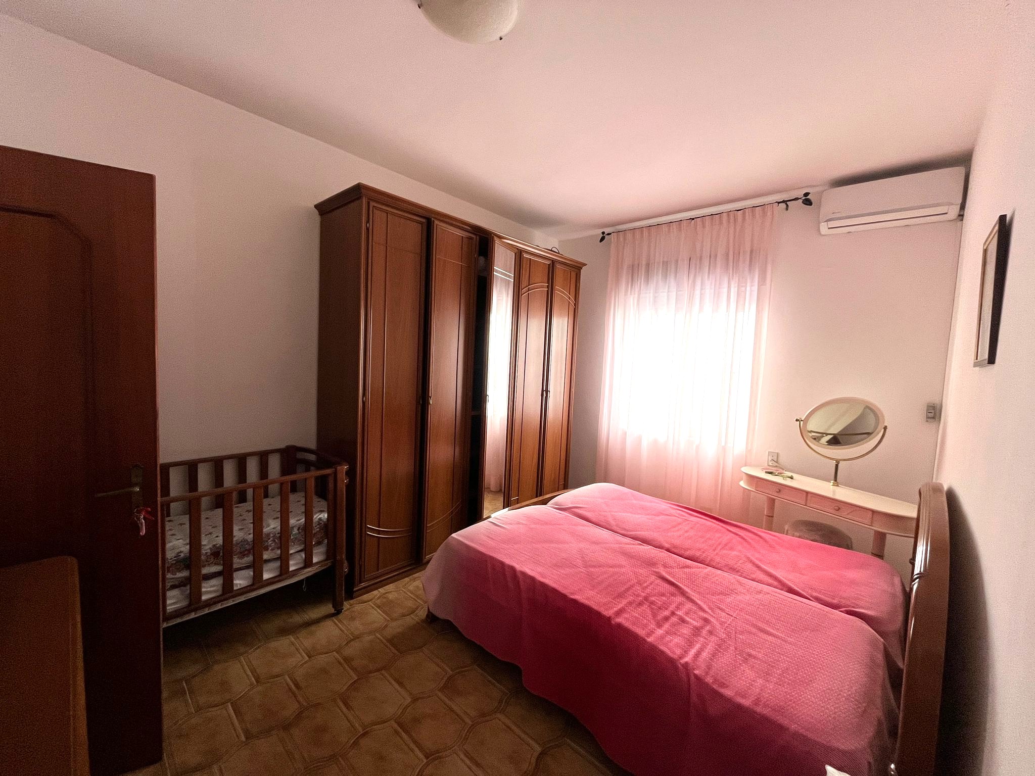 Letto matrimoniale con lenzuola rosa in camera da letto, armadio in legno, culla per neonato e specchio da toeletta, ambiente luminoso e accogliente, ideale per soggiorno confortevole e funzionale a NextCasa.