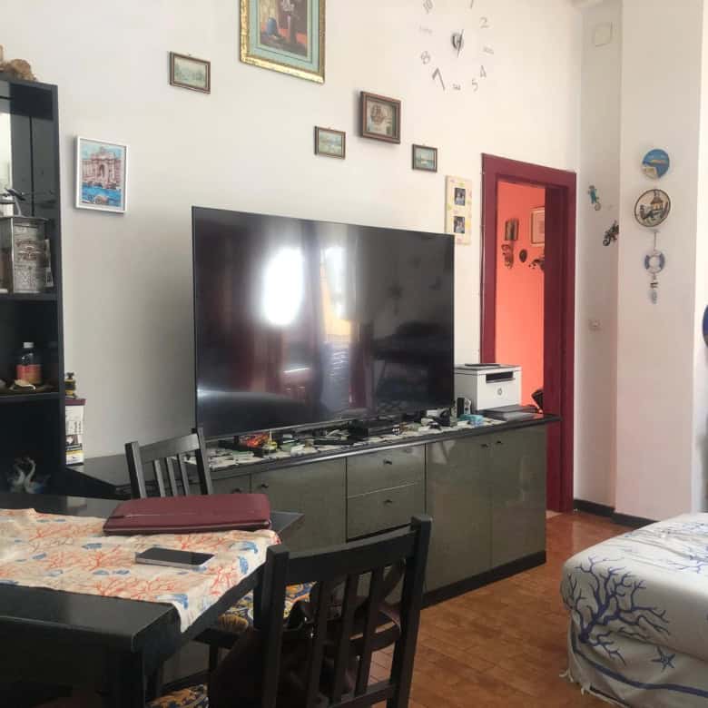 Parete soggiorno decorata con quadri e orologio grande, TV sopra mobile basso con vari oggetti, area pranzo con tavolo e sedie, arredamento e oggetti decorativi portano a uno stile conviviale e accogliente.