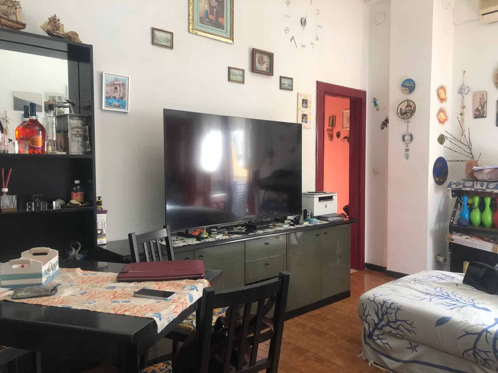 Parete soggiorno decorata con quadri e orologio grande, TV sopra mobile basso con vari oggetti, area pranzo con tavolo e sedie, arredamento e oggetti decorativi portano a uno stile conviviale e accogliente.