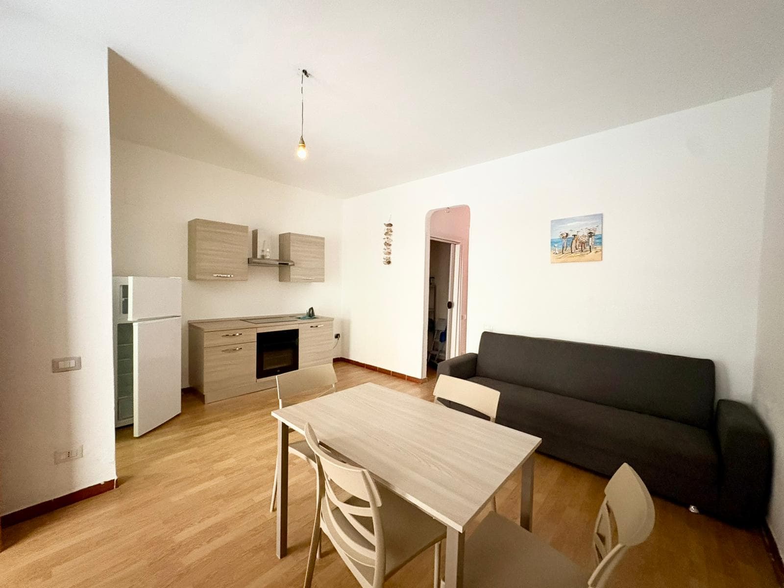 Interno di un appartamento con soggiorno e cucina open space, arredato in stile minimalista con mobili chiari e divano nero, pavimento in legno e pareti bianche.