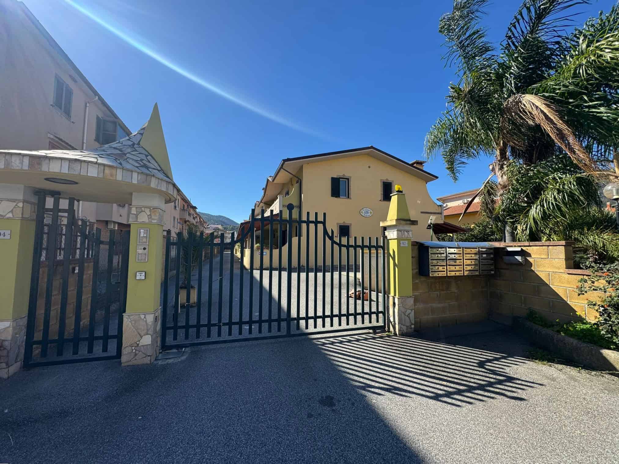 Casa privata con cancello in ferro battuto e giardino tropicale, situata in una zona residenziale soleggiata con palme e piante esotiche, ideale per famiglie e investimenti immobiliari.
