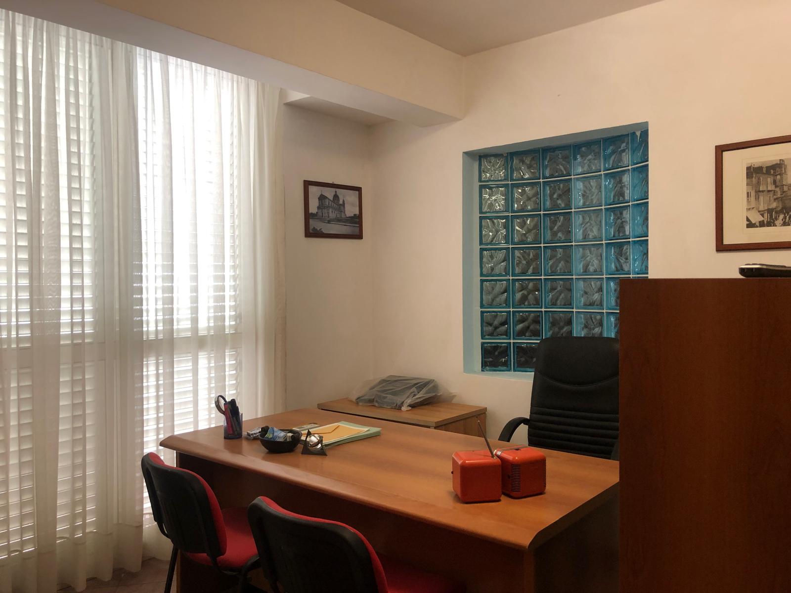 Ampia scrivania in ufficio con sedia ergonomica, oggetti da lavoro e decorazioni, illuminata da luce naturale attraverso tende trasparenti, ambiente professionale e accogliente.