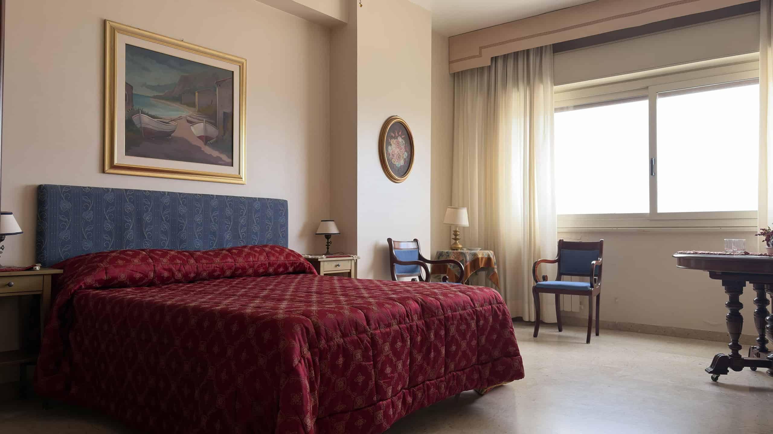 Letto matrimoniale con testiera imbottita blu in una camera da letto luminosa e accogliente, con tende chiare e mobili classici.