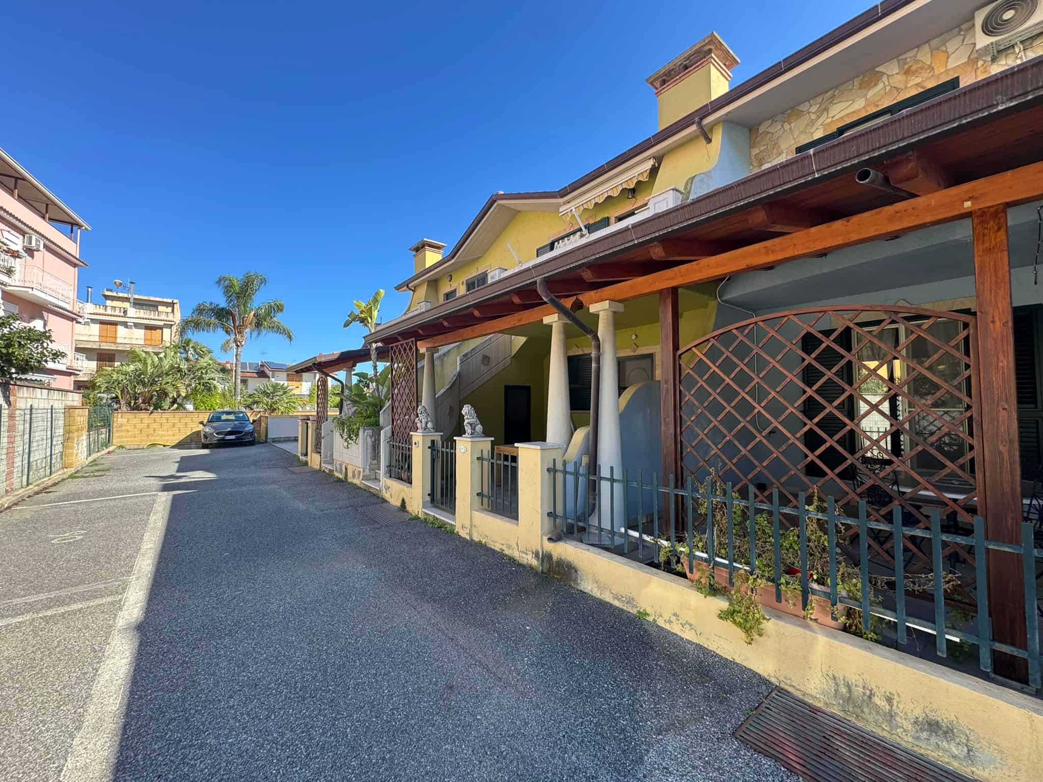 Ampia strada residenziale con case colorate e giardini ben curati, sotto un cielo azzurro e soleggiato, in un quartiere tranquillo e ben servito.
