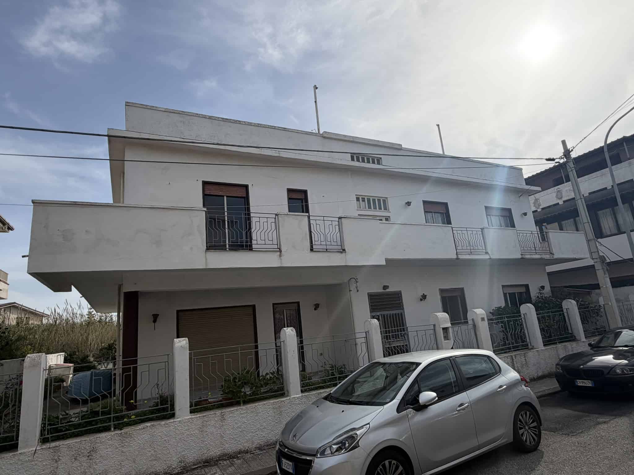 Edificio residenziale moderno a due piani con balconi e garage, situato in zona urbana, con auto parcheggiate davanti e cielo sereno.
