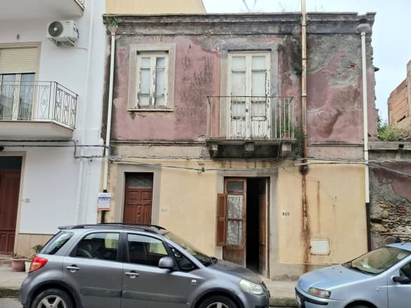 Antico edificio residenziale in centro città con pareti esterne in pietra e segni di deterioramento, situato tra edifici moderni, con auto parcheggiate davanti.