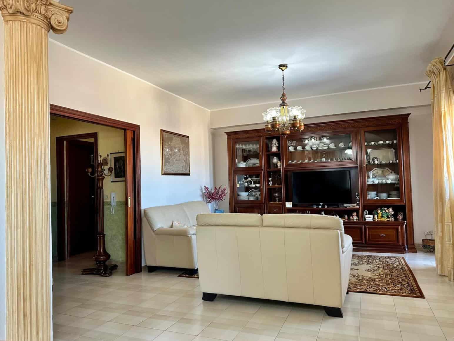 Ampia zona living con divani bianchi, mobile porta TV in legno scuro e lampadario elegante, ideale per soggiorni confortevoli e raffinati.