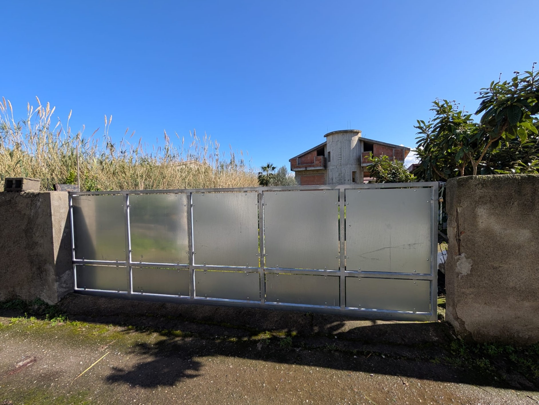 Cancello in metallo con pannelli opachi, installato tra muri in cemento, in un ambiente esterno soleggiato con cielo azzurro.