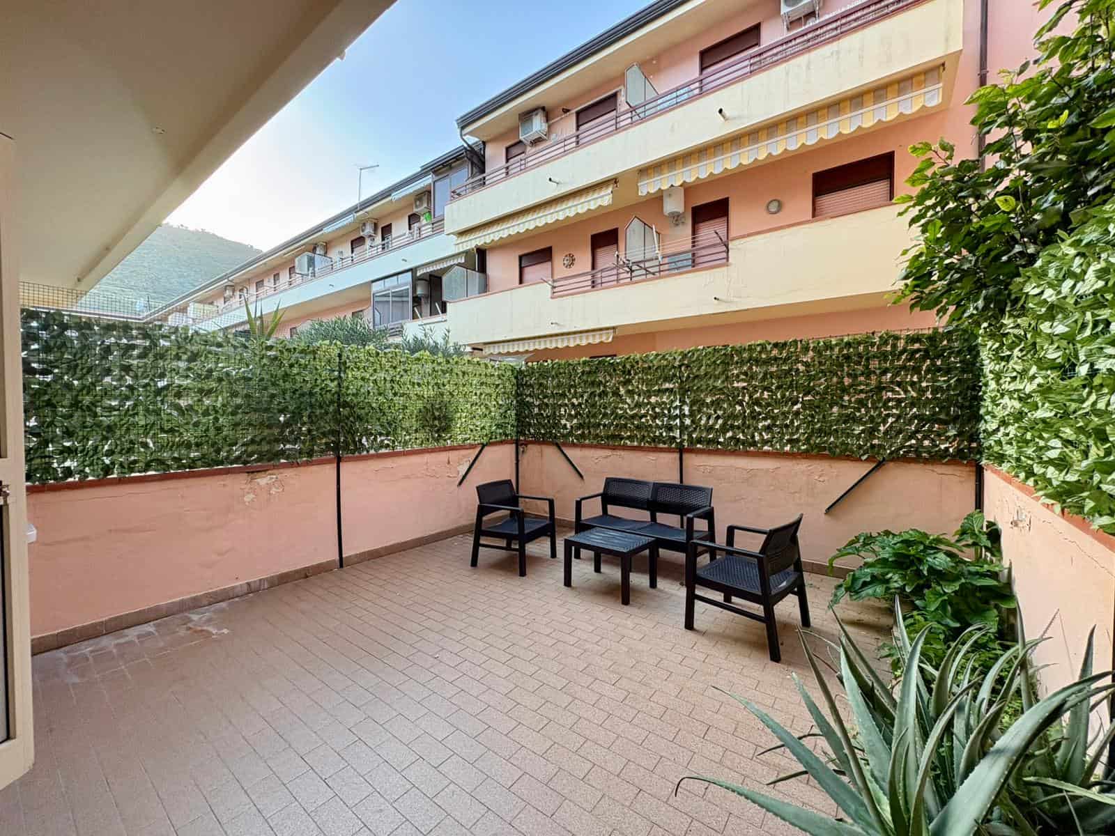 Balcone spazioso con vista su complesso residenziale, arredato con sedie e tavolino, circondato da piante verdi, ideale per relax e momenti all'aperto in un contesto urbano tranquillo.