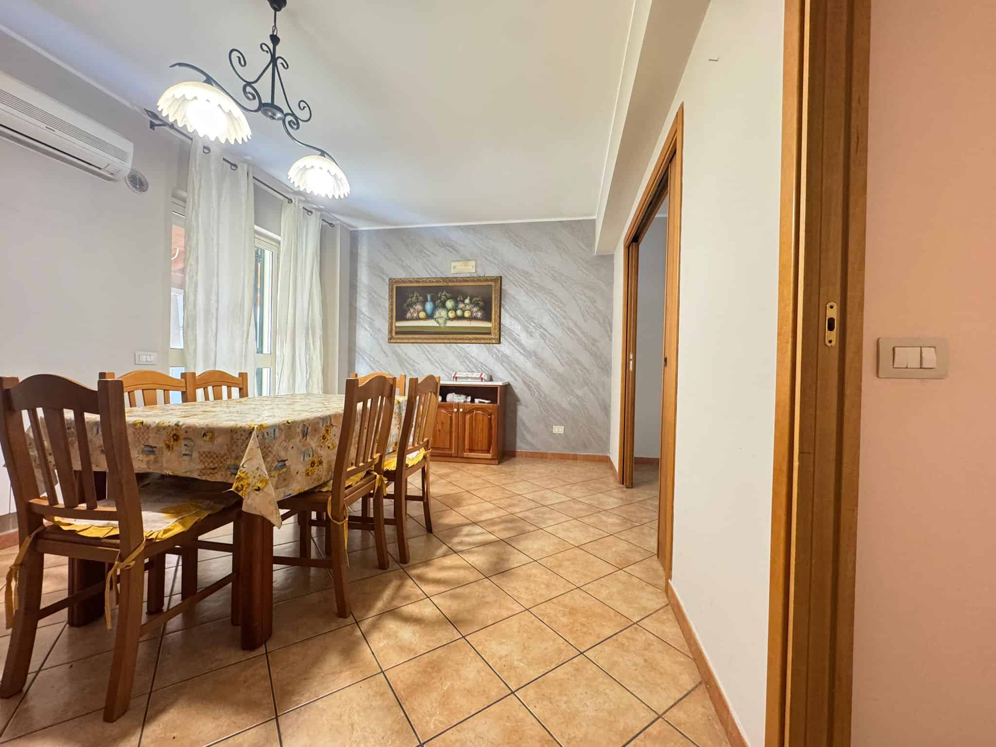 Ampia sala da pranzo con tavolo in legno e sedie, illuminata da lampadari eleganti, in un ambiente luminoso e accogliente con pareti decorate e grandi finestre con tende bianche.