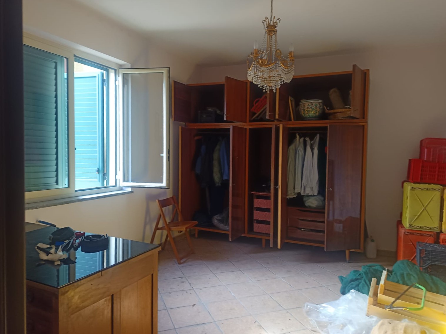 Armadio a muro in legno con ante aperte, ripostiglio e spazio per vestiti e accessori, in una stanza luminosa con finestra aperta e lampadario elegante.