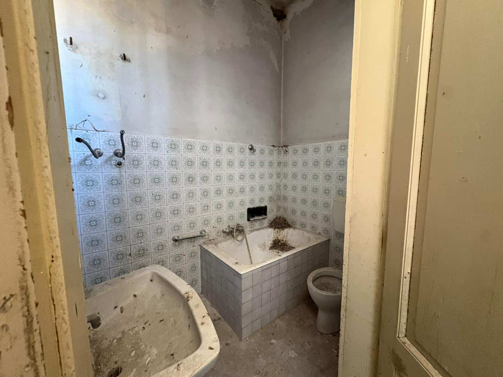 Il bagno in condizioni di abbandono con piastrelle vecchie e sporche, vasca da bagno rovinata e wc sporco, ambiente trascurato e in cattivo stato di manutenzione.