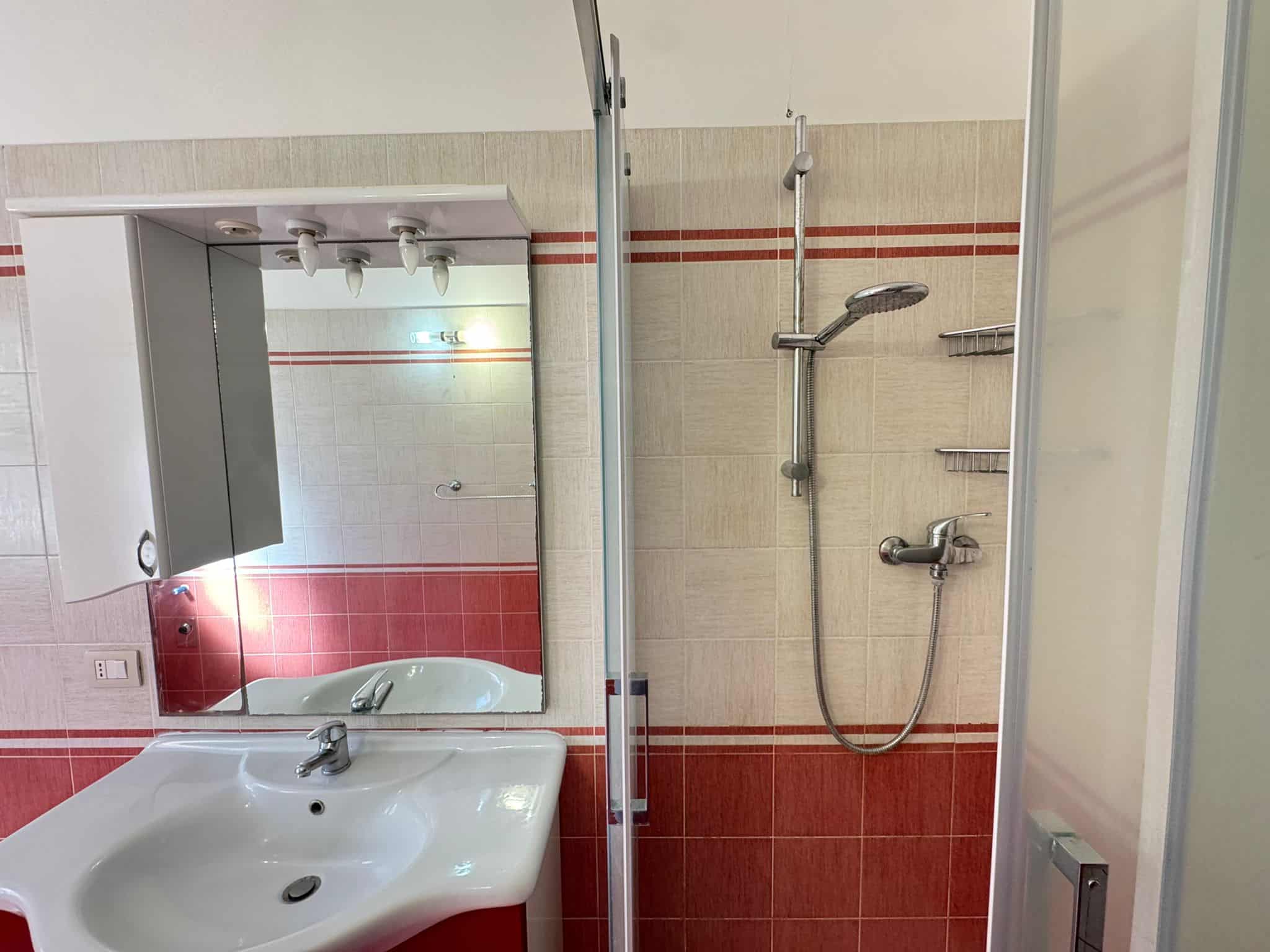 Lavabo e doccia in un bagno moderno con piastrelle beige e rosse, specchio e accessori essenziali, ideale per appartamenti in vendita o affitto a NextCasa.