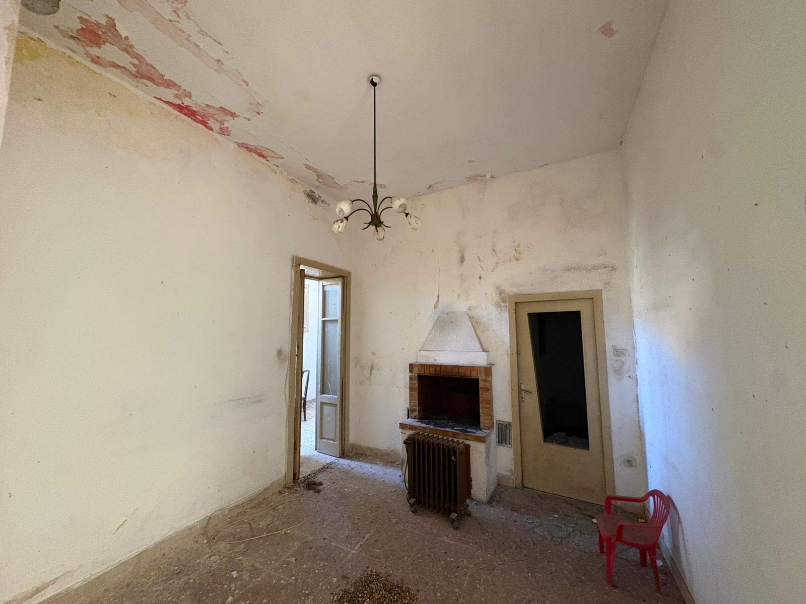 Vecchio soggiorno con camino in mattoni e pareti in cattive condizioni, pronto per ristrutturazione, con porta e finestra che portano luce naturale, e sedile rosso in plastica vicino al camino.