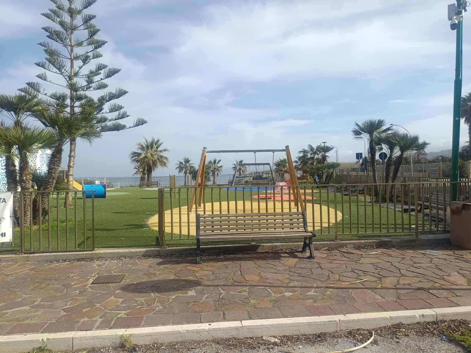 Panchina e area giochi all'aperto vicino al mare con palme e vegetazione tropicale, ambiente rilassante e ideale per famiglie e bambini, vista panoramica e atmosfera estiva.