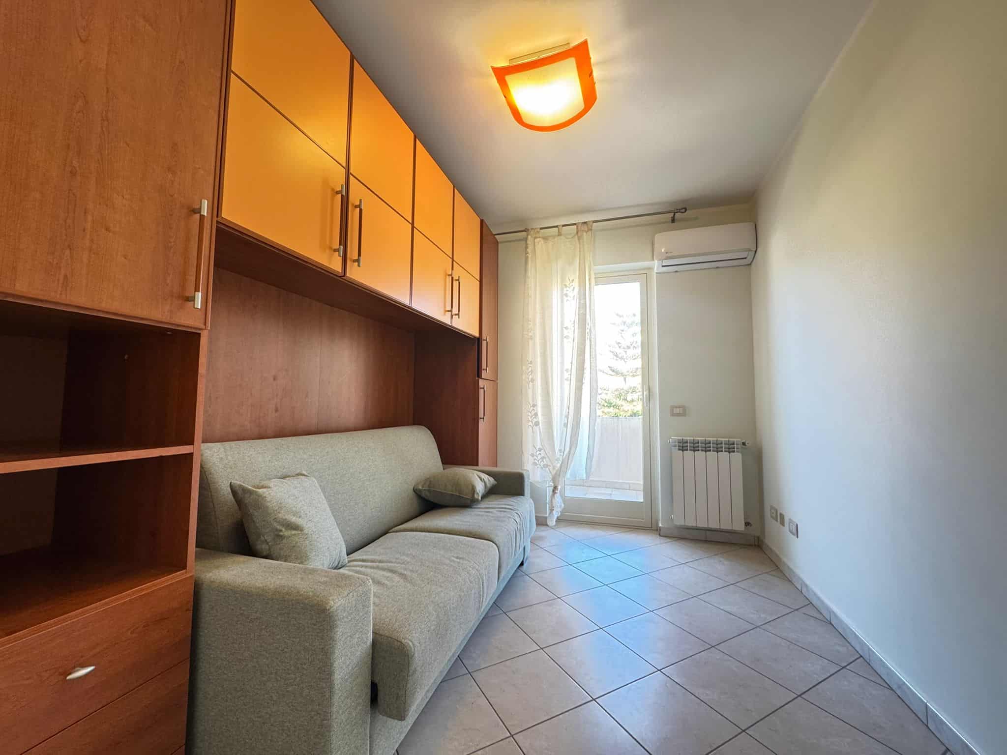Soggiorno luminoso con mobili in legno e divano beige, finestra con tende leggere e aria condizionata, ideale per affitti brevi o residenziali a NextCasa.