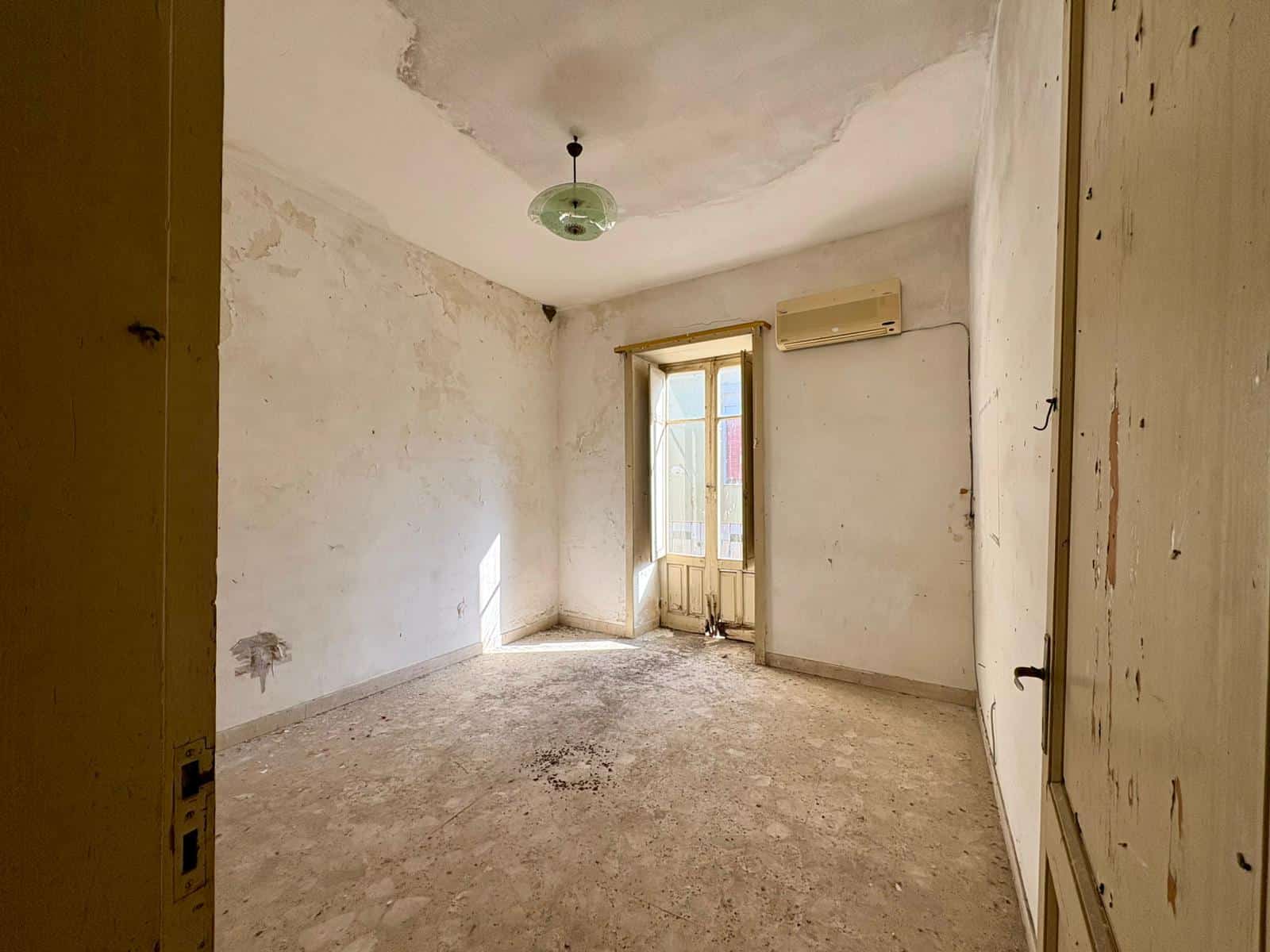 Stanza vuota in fase di ristrutturazione con pareti bianche, pavimento in cemento e finestra che lascia entrare luce naturale, ideale per rinnovare e personalizzare la casa.